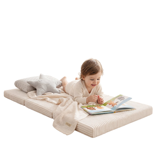 Lifestyle-Bild: Kleinkind in cremefarbenem Strickpulli liegt bäuchlings auf der ausgeklappten Reisebettmatratze in Beige und blättert lächelnd in einem Bilderbuch, daneben ein Plüsch-Stern und eine cremefarbene Kuscheldecke.