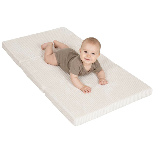 Baby in beigefarbenem Body liegt lächelnd auf der ausgeklappten Reisebettmatratze in Beige mit Cord-Bezug, Blick in die Kamera.