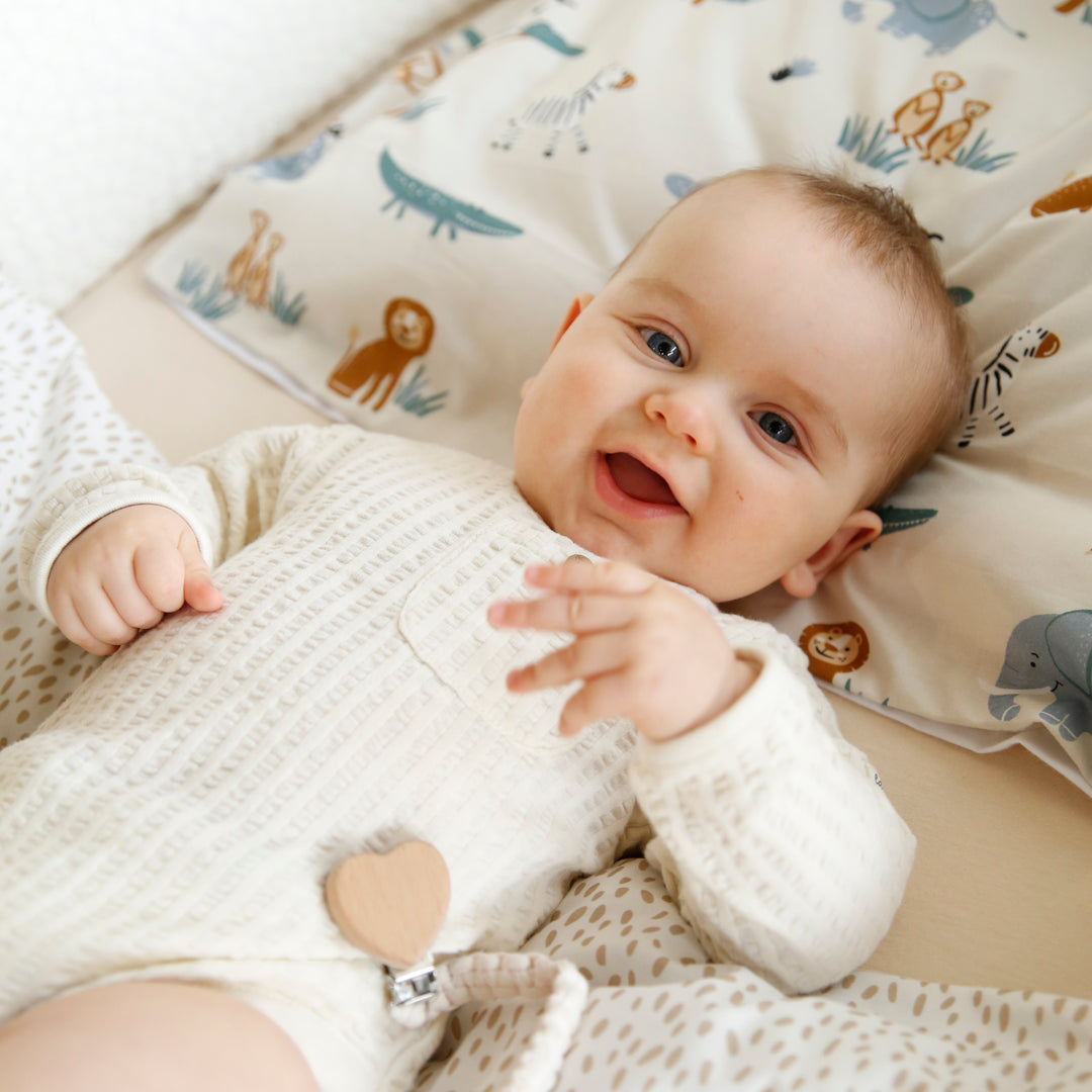 Bébé riant en barboteuse tricotée allongé sur le linge de lit "Savanna Friends" avec des motifs animaliers en arrière-plan