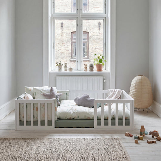 Lit bas Montessori laqué blanc près d'une fenêtre haute, avec lampe en papier et coussins étoile et nuage. Partiellement généré par IA