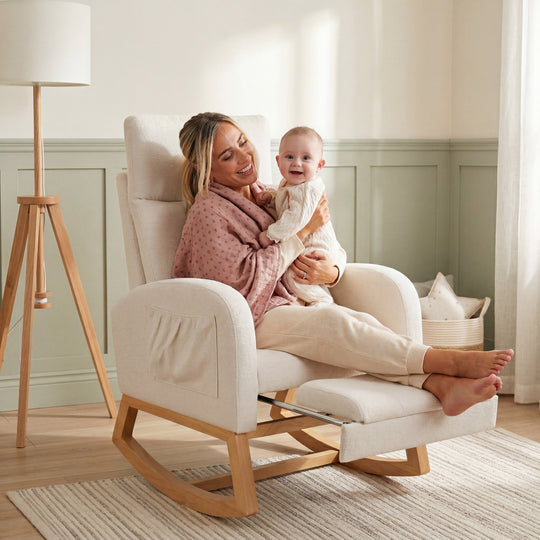 Lifestyle-Bild: Eine lächelnde, barfüßige Frau in rosa Strickjacke sitzt mit einem lachenden Baby im Arm im Relaxsessel in Beige mit ausgezogener Fußstütze, im Hintergrund ein Kinderzimmer mit salbeigrüner Wandvertäfelung und Stehlampe.
