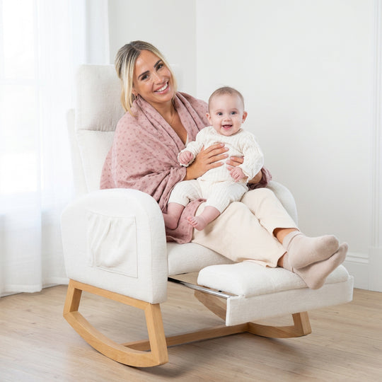 Lifestyle-Bild: Lächelnde Frau mit rosa Tuch hält ein lachendes Baby im Relaxsessel in Beige mit ausgezogener Fußstütze und Schaukelkufen aus Holz in einem hellen Zimmer.