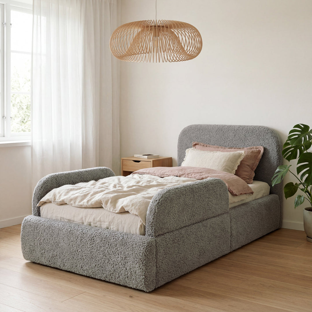 Cama infantil gris de bouclé con protección anticaídas en una habitación luminosa con lámpara de ratán y planta. Generada íntegramente con IA.