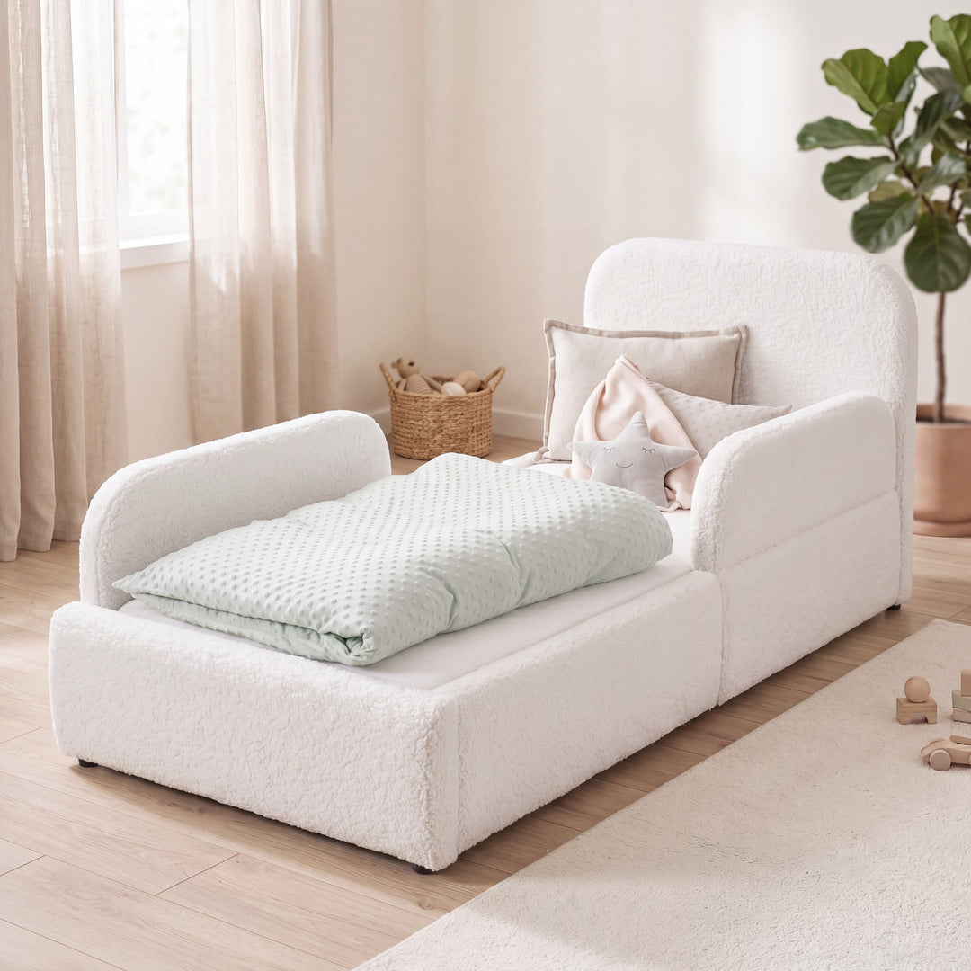 Cama infantil blanca de bouclé con protección anticaídas, cesta de ratán y planta en una habitación luminosa. Generada íntegramente con IA.