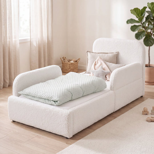 Cama infantil blanca de bouclé con protección anticaídas, cesta de ratán y planta en una habitación luminosa. Generada íntegramente con IA.