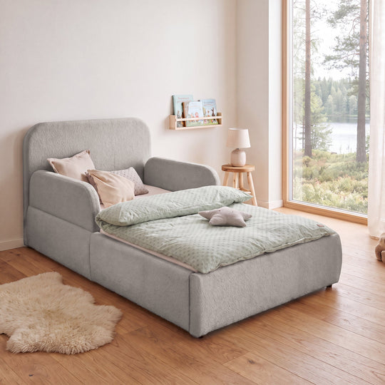 Cama infantil roba Little Dream gris claro con protección lateral en una habitación infantil con vistas al lago y lámpara de pie. Generada parcialmente con IA.