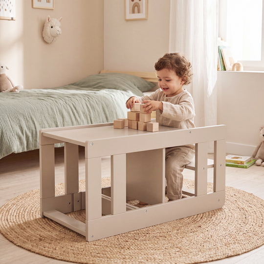 Toddler sits on the converted seating set of the Cashmere learning tower and plays with wooden building blocks, in the background a bed with green bedding and a round jute rug.